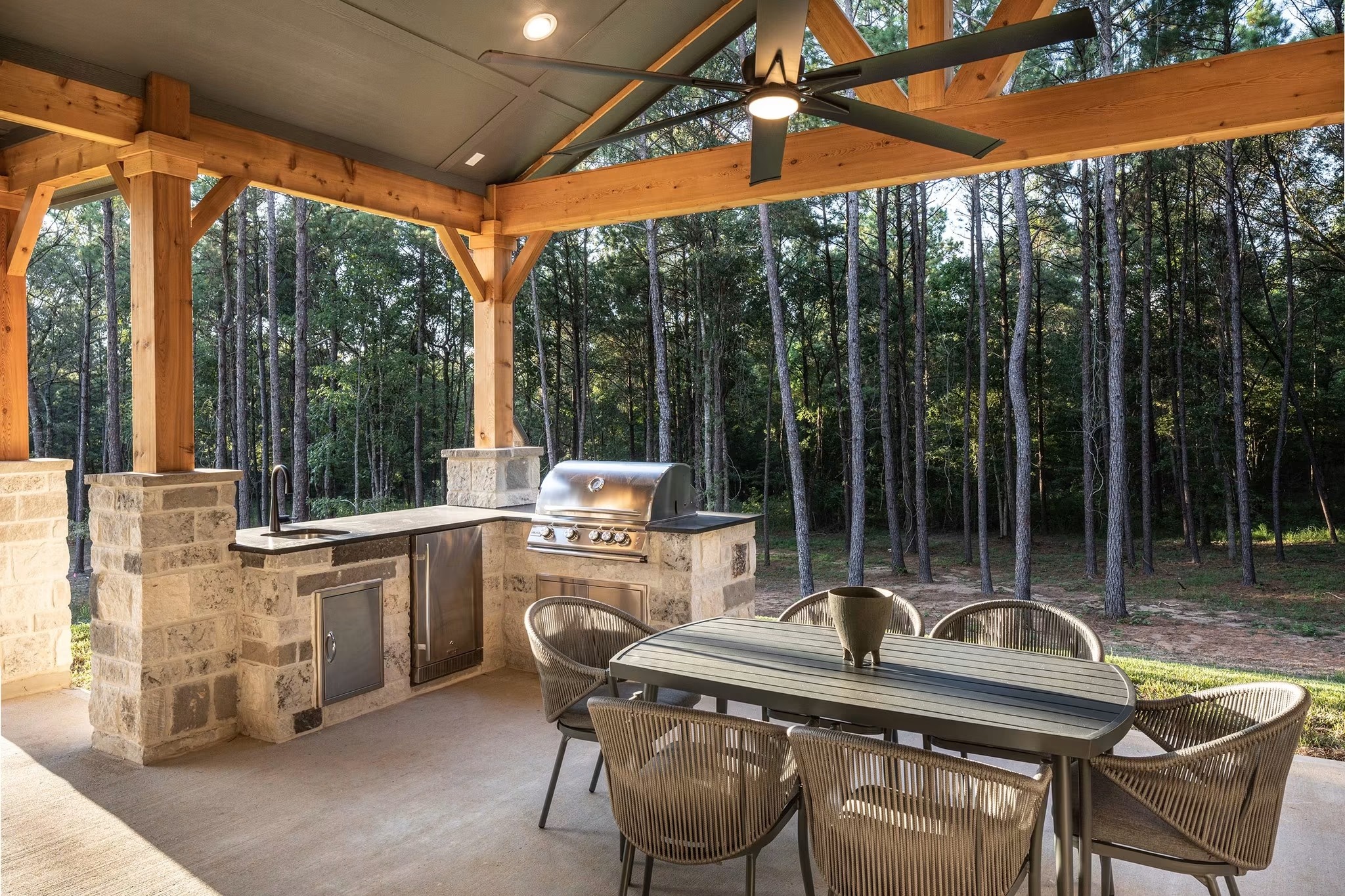 Covered outdoor kitchen with stone counters and dining table overlooking tall pines